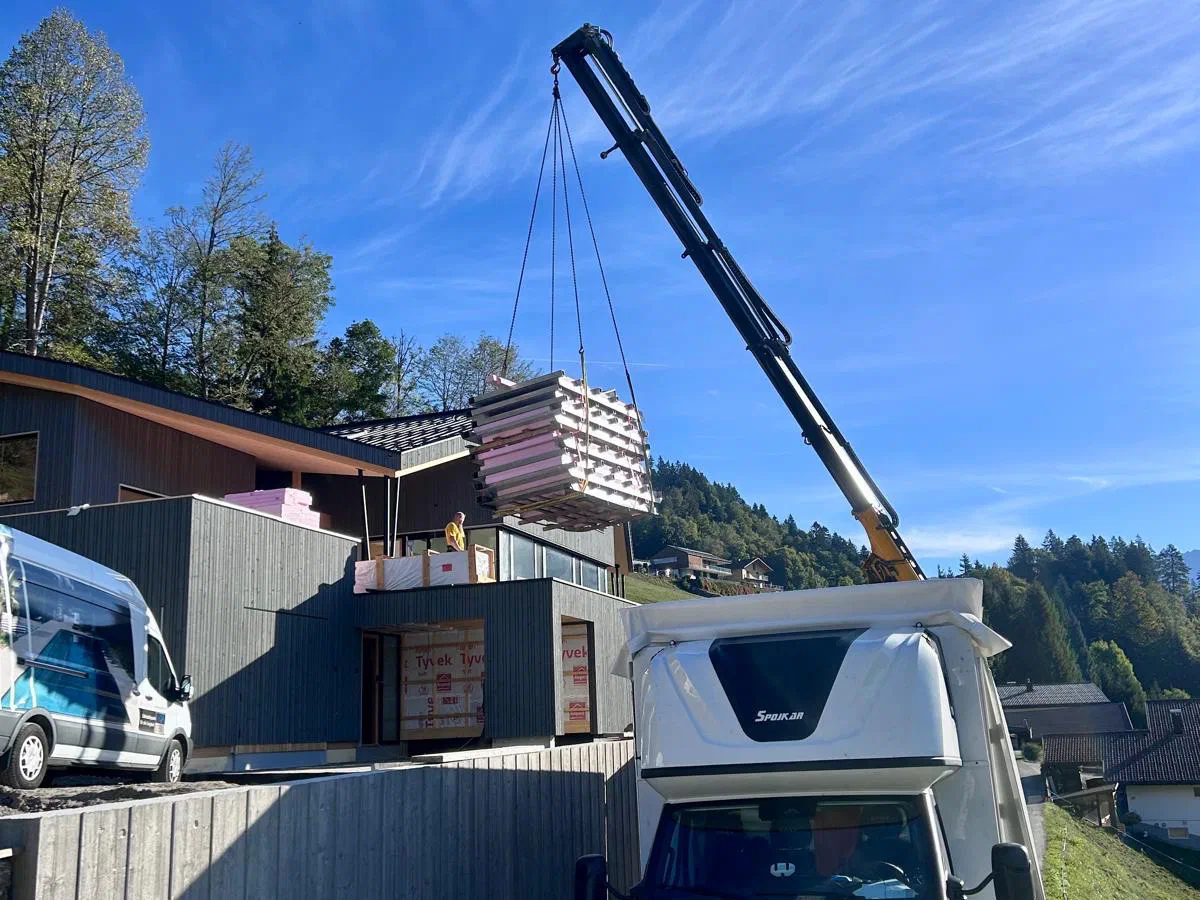 Ein Kran hebt Ladung durch das geöffnete Dach eines Planenfahrzeugs von DMF Express auf einer Baustelle in den Alpen